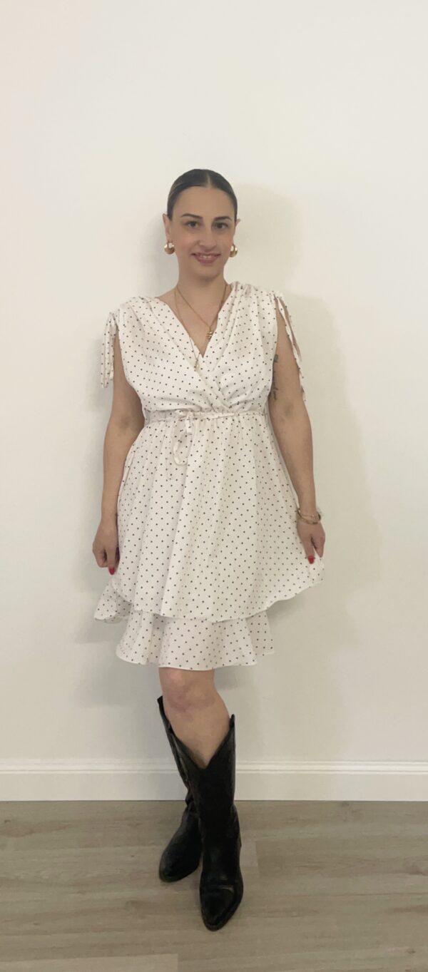 Abito corto bianco con pois neri, scollo incrociato, spalline da annodare e balza sul fondo. Indossato da modella alta 1,60 m, taglia S, con stivali texani neri.