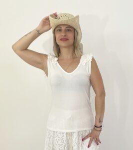 Canotta bianca senza maniche con scollo a V e rouche sulle spalle, indossata da modella con cappello di paglia.
