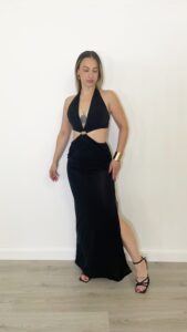 Donna indossa abito lungo nero con cut-out laterali, scollo allacciato dietro il collo e dettaglio gioiello centrale.