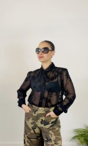 Camicia nera trasparente con ricami floreali e fiocco al collo, indossata con pantaloni mimetici.