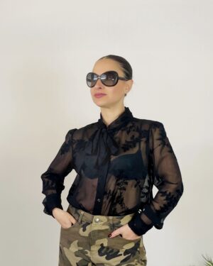 Camicia nera trasparente con ricami floreali e fiocco al collo, indossata con pantaloni mimetici.